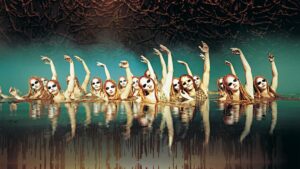 Several red haired dancers with white masks on in a pool of water with one arm raised